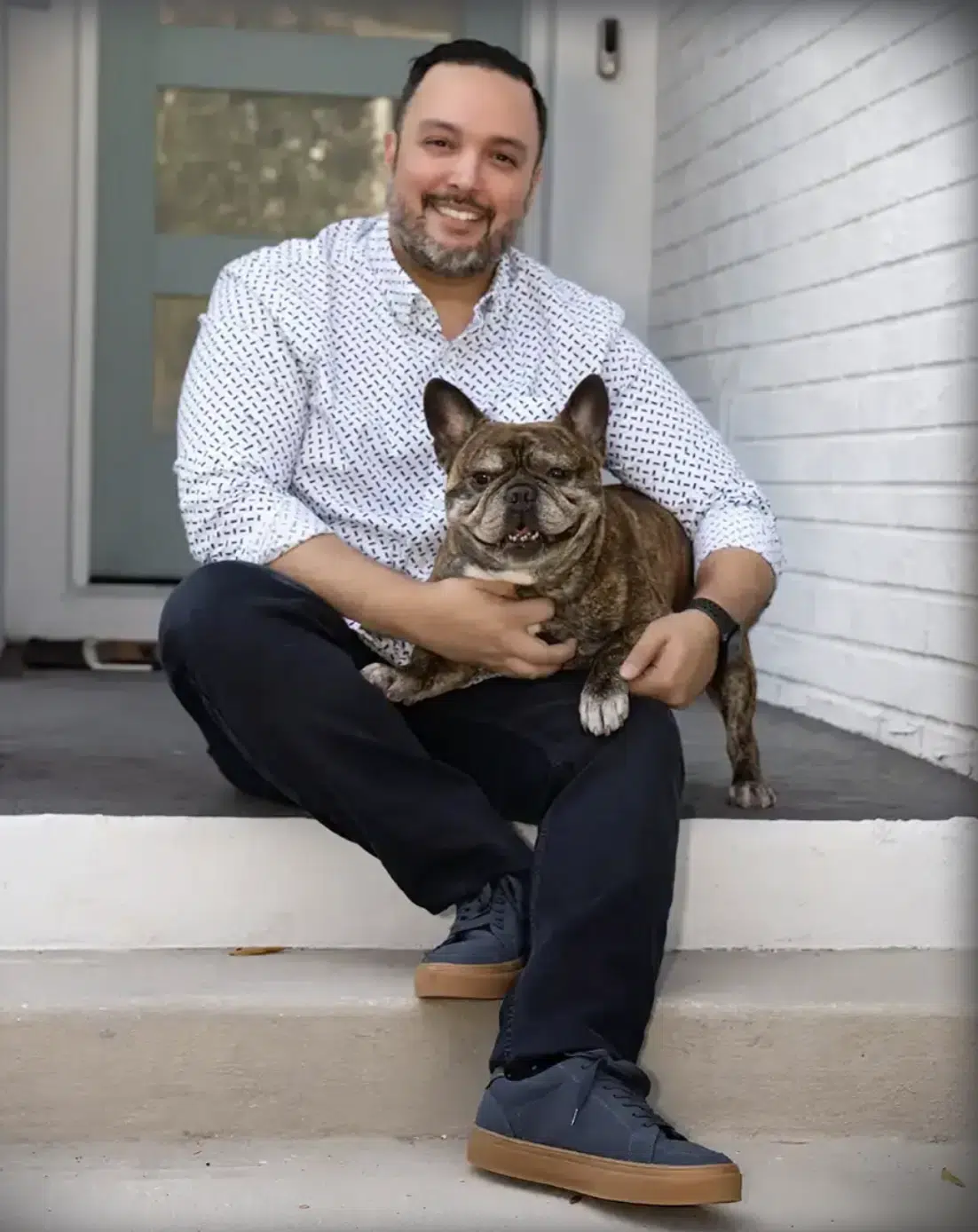Un hombre con barba está sentado en el escalón de un porche, sonriendo, mientras sostiene en su regazo a su bulldog francés atigrado, capturando un momento perfecto de cierre de año.